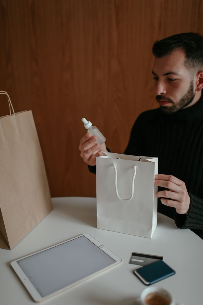 Services Crop self assured young bearded male buyer in casual clothes sitting at table and checking delivered purchases made via tablet with credit card at home
